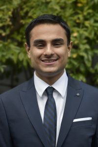 Karan Khosla indian guy in navy blue suit with blue tie with black strips and white shirt and green tree in background
